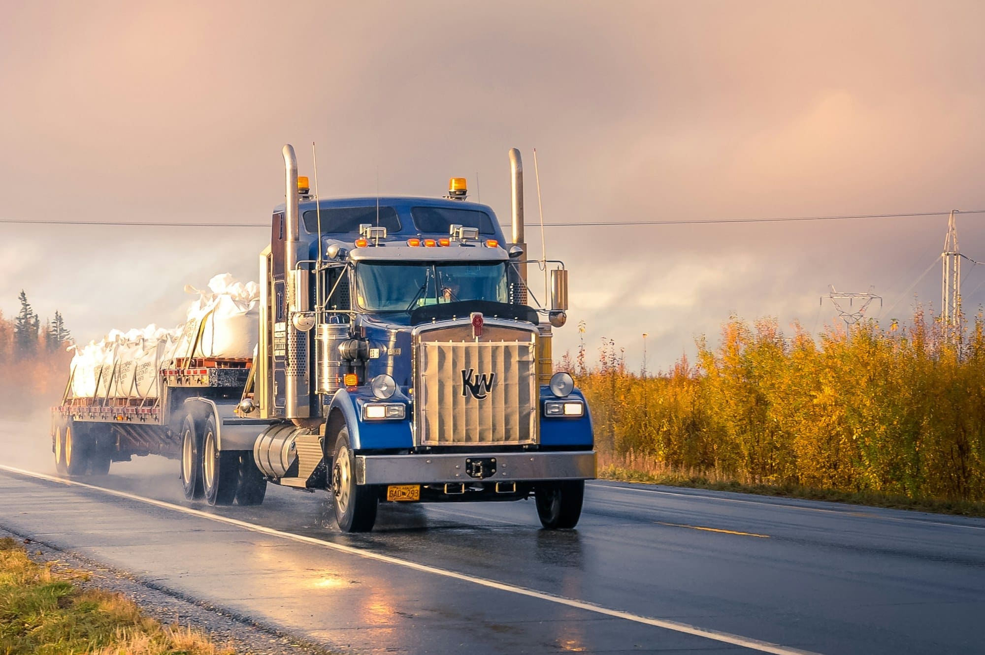 A conventional semi tractor pulling a covered trailer down a wet two-lane highway under a clearing sky.